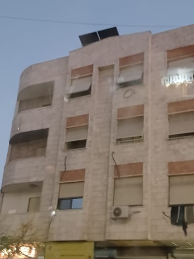The image depicts the exterior of a multi-story building in Kafr Sousa Municipality, Syria, viewed from a slightly upward angle against a twilight sky. The building features a light-colored, possibly stone or concrete facade with a rounded corner on its left side.
On the roof, two dark, rectangular solar panels are visible. The building has multiple vertical rows of windows, most equipped with roll-down shutters. Several shutters are partially raised, revealing interior spaces or windows behind them. Specifically, shutters on the upper-left, middle-left (second from left), and upper-right windows are partially open. One window on the middle-left, beneath a partially raised shutter, displays a blue-tinted pane. A reflection of light-colored text, possibly Arabic characters, is visible on a window pane on the upper-right side.
Two white, rectangular outdoor air conditioning units are mounted on the facade. One is located on the lowest visible floor, second from the right, beneath a window. Another is situated on the far right of the lowest visible floor, below which dark fabric items, possibly laundry, are hanging. Dark, insulated electrical wires run vertically down the facade in several sections. A circular marking is present on the facade between windows on the middle floor, second from the left. A small, downward-pointing light fixture is visible on the middle-left section.
Below the main facade, a blurred yellow sign is partially visible at ground level, suggesting a commercial establishment. Blurred tree branches or foliage are also visible on the lower-left. The sky above is a clear, light blue to grey, indicative of dawn or dusk. No individuals are visible in the scene.