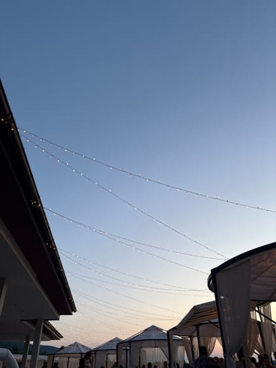 The image depicts an outdoor scene with a clear sky at dusk. Several strings of small, warm-toned lights are strung across the sky, creating a festive ambiance. In the foreground, the edge of a building with a dark brown roof is visible on the left. Beyond that, there are several white gazebos with draped curtains, suggesting an outdoor dining or event space. Some people are visible in and around the gazebos, engaged in what appears to be a gathering. The overall setting evokes a tranquil evening atmosphere. The location is Skalsko, Bulgaria.