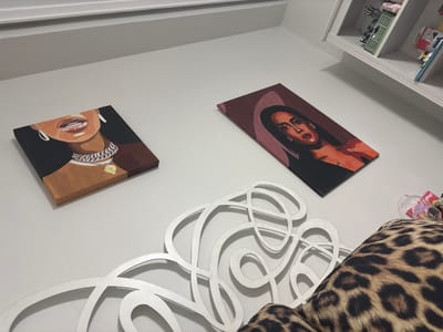 The image shows a section of a white wall adorned with two rectangular canvas paintings. The painting on the left depicts a partial view of a person's face, focusing on the mouth, neck, and jewelry. The one on the right shows a full portrait of a person with dark hair. A decorative white metal headboard with swirling patterns is mounted below the paintings. A leopard print cushion is visible to the right of the headboard. The picture was not taken in Córdoba, Spain.