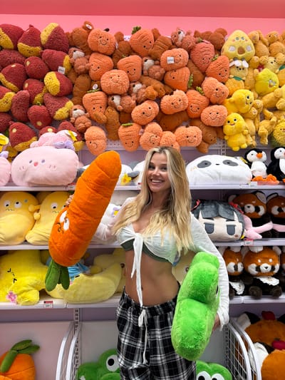 The image shows a young woman with blonde hair, smiling and looking at the camera, as she is surrounded by a large collection of plush toys. The plush toys are primarily in shades of orange, yellow, and brown, with some pink and green accents. There are stuffed carrots, pumpkins, and teddy bear-like creatures, among others. Many of the toys have embroidered or appliquéd facial features.
The woman is wearing a light-colored top and black and white plaid pants. She is positioned amongst the toys, with some of them obscuring parts of her body. In the background, more plush toys are visible on shelves, suggesting a retail environment or a display. The setting appears to be indoors, possibly a store.
The woman's expression is happy and engaged. The overall tone of the image is cheerful and playful, likely due to the abundance of cute stuffed animals and the woman's smile. No specific time of day or weather conditions are discernible. There is some text visible on the price tags of the plush toys, such as "149" and "359", indicating prices in Thai Baht, which is consistent with the specified location context of Patong, Thailand. The image is a portrait of the woman interacting with a fun and colorful assortment of merchandise.