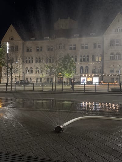 The image depicts a scene at night on a street in Poznan, Poland, likely near the historic Poznan City Hall or a similar grand building. In the foreground, a flexible white hose is on the wet, paved ground, spraying water in a fan shape. The pavement is tiled and glistening from the water, with reflections of lights from the street and buildings visible on its surface. Beyond the immediate foreground, a metal railing separates the paved area from a street or tram line. Across the street, a large, imposing building with multiple windows and an arched facade is illuminated. The building appears to be made of stone and has a dark roof. A sign on the building reads "BANK POCZTOWY SA". Further down the street, a tram stop with a sheltered waiting area is visible, with a few people sitting on benches and one person standing. The sky is dark, and there is a misty or smoky haze in the air, particularly around the upper floors of the building. A dark-colored car is parked on the street in the middle ground. The overall atmosphere is nocturnal and slightly dramatic due to the lighting and the spray of water.