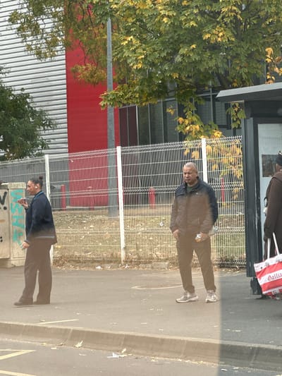 The image displays an outdoor urban scene in Stains, France, under overcast daylight conditions, featuring autumnal foliage. Three individuals are present on a paved sidewalk or street edge.
On the left, a person, appearing female, with dark hair pulled back, stands in profile, facing right. She wears a dark blue, long-sleeved top with a light stripe and dark trousers. A white earbud is visible in her right ear, and she holds a small, light-colored object to her mouth.
In the center, a middle-aged man with short, graying hair stands facing slightly left. He is dressed in a dark quilted jacket, a dark shirt, dark trousers, and light-colored athletic shoes. He holds a crumpled, light-colored object in his left hand.
To the far right, partially obscured by a dark-framed bus stop shelter, another person, wearing a dark brown coat and a dark head covering, is visible from the chest up, holding a large red and white shopping bag with "Gallia" printed on it.
The immediate foreground consists of asphalt pavement and a low curb, scattered with fallen leaves and small pieces of litter. Behind the individuals, a continuous white metal mesh fence separates the paved area from a strip of dry grass. Beyond the fence, a large building with sections of grey corrugated metal siding and red flat panels, along with windows, is visible. Green trees with yellowing leaves partially obscure the building and a grey utility pole. A concrete block with faded graffiti is present behind the leftmost individual.
