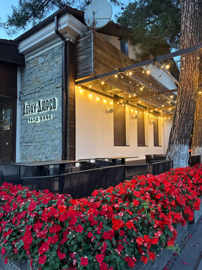 The image depicts the exterior of the "Abrau-Dyurso Grand Cafe" located in the resort town of Abrau-Dyurso, Russia. The cafe features a stone facade with a wooden upper section. An illuminated sign with the cafe's name is prominently displayed on the stone wall. Outdoor seating is arranged with dark-colored tables and chairs, separated by black fencing. A lush planter box filled with vibrant red flowers runs along the front of the seating area. String lights are strung across the outdoor patio, illuminating the space. Large trees with textured bark are visible to the right of the building. The sky above is a dim blue, suggesting it is either early morning or late evening. No people are visible in the image.