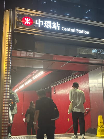 The image depicts the Central Station in Hong Kong. In the foreground, a sign displays the station's name in both English and Chinese, with the MTR logo visible below. The station platform is visible, where several people are walking or standing. A yellow caution sign stands near the platform edge. The lighting appears to be a mix of natural and artificial. The walls and floors are predominantly red.
