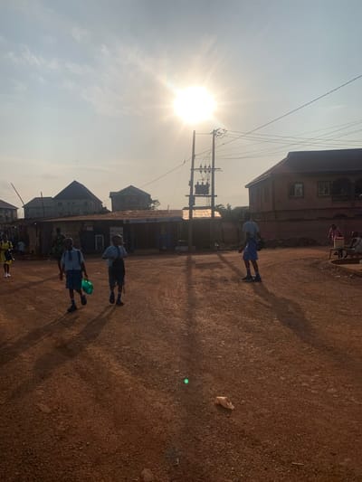 This is a wide shot of an outdoor scene in Enugu, Nigeria, depicting a street with several people and buildings under a bright, hazy sun. The foreground is a dirt road, with two young boys in school uniforms walking from left to right, carrying backpacks and a green bag. They are casting long shadows. In the middle ground, there are several structures resembling market stalls or small shops made of corrugated metal. Behind these structures, larger buildings are visible, including a two-story house with a reddish-brown facade. A utility pole with a transformer and electrical wires is prominent in the mid-ground. A few other figures are scattered in the background, including a woman in a yellow dress on the far left and a person sitting near the house on the right. The overall impression is of a dusty, sun-drenched street scene in a residential or commercial area.