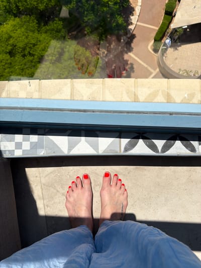 A first-person perspective shot shows bare feet with bright red toenail polish resting on a light-colored tiled floor bathed in strong sunlight. The feet are positioned near the edge of a balcony or outdoor area. Beyond the feet, a blue frame of a window or door is visible, with decorative patterned tiles below it. Through the glass, a reflection of the feet and the surrounding greenery, including trees, is visible. A glimpse of an outdoor paved area with a curved wall and some landscaping can also be seen in the reflection, suggesting an urban setting. The overall impression is one of leisure and relaxation, possibly on a sunny day in Ashkelon, Israel.