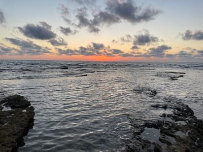 The image captures a sunset over the Mediterranean Sea, as seen from the rocky coast of Haifa, Israel. The scene is divided horizontally by the horizon. The upper portion of the image depicts the sea and the rocky shore. The water is slightly choppy, reflecting the dimming light of the sky. Dark, textured rocks jut out from the left and right sides, framing the view of the water. In the lower portion, the sky transitions from a warm, soft orange glow near the horizon, indicative of the setting sun, to a pale, grayish-blue at the top. Scattered, dark clouds drift across the sky, their undersides catching the last rays of sunlight. The overall mood is serene and peaceful, with the natural beauty of the coastal landscape at dusk. There is no visible text in the image.