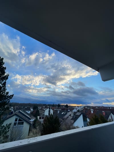 The image displays an elevated view of a residential area in Senden, Germany, observed from a balcony. The upper portion of the frame is obscured by a dark grey concrete balcony ceiling, while a light grey concrete railing is present at the bottom, partially framing the scene. The sky features a mix of deep blue and numerous white to grey cumulus clouds. A soft orange-yellow glow is visible on the distant horizon to the right, indicating either sunrise or sunset. Below the horizon line, a dense band of trees and foliage forms a distant perimeter. The middle ground and foreground consist of a dense arrangement of detached and semi-detached houses. These structures exhibit varied architectural styles, primarily featuring pitched roofs with red-brown or dark grey tiles and light-colored facades. Some residences show modern elements like large angular windows and rooftop solar panels. Interspersed among the houses are various trees and bushes, including both evergreens and deciduous trees with bare branches. A white passenger vehicle is visible on a street between houses in the mid-left area of the frame. No individuals are present in the scene.