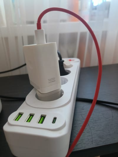 A white power strip with multiple outlets is placed on a dark wooden surface. Three green USB ports are visible on the front of the power strip. A white power adapter labeled "160W SUPERVOOC" is plugged into one of the outlets. A red cable is connected to the power adapter and extends upwards and to the right. In the background, sheer white curtains hang in front of what appears to be a window. The scene suggests an indoor environment, possibly a room in Kalynivka, Ukraine. There are no people visible in the image. The lighting is moderate, with some light coming from the background.