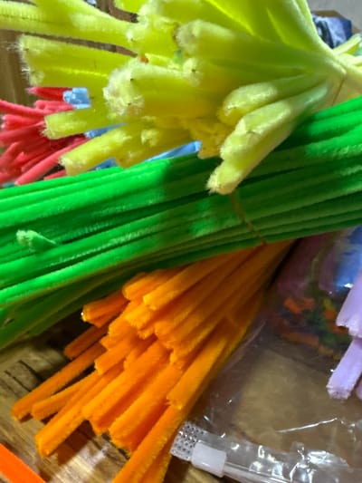 The image presents a close-up view of several bundles of pipe cleaners (chenille stems) in various colors. Dominant colors include yellow, green, orange, and red. The bundles appear to be tightly packed together. A clear plastic bag containing more pipe cleaners of assorted colors is partially visible in the background. A plastic container is visible at the right of the image. The environment appears to be indoors on a wooden or laminate surface. There are no visible indications of the location being in Troyan, Bulgaria.