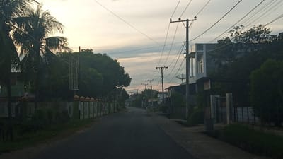 The image depicts a quiet, paved road in Meunasahteungkudigadong, Indonesia, during what appears to be early morning or late evening, indicated by the soft, diffused light and overcast sky. The road is lined on both sides with vegetation, including palm trees on the left and denser trees on the right. Structures are visible along the road, including houses and fences. On the left, a green-roofed building with white fencing and decorative pillars topped with golden domes is present. On the right, a modern, two-story white building with dark trim stands. Numerous utility poles with complex networks of wires crisscross the sky, typical of urban or suburban environments. A satellite dish is visible on the roof of a building on the right. The overall atmosphere is serene and unremarkable, suggesting a typical residential street scene in this Indonesian locale. No people are visible in the frame.