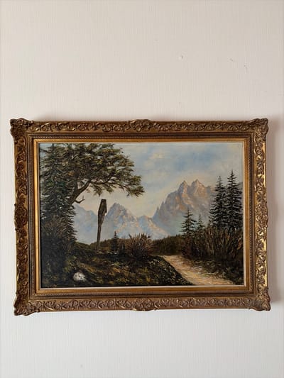 The image shows an oil painting of a mountain landscape. The painting is framed in an ornate gold-colored frame. The scene depicts a dirt path leading through a wooded area towards a range of majestic mountains. A wooden signpost stands on the left side of the path. In the foreground, there are trees and shrubs, and in the background, the mountains are painted with soft, cloudy skies. The painting is hanging on a textured white wall, which appears to be wallpaper. There is no information within the image to indicate its location in Osnabrück, Germany.