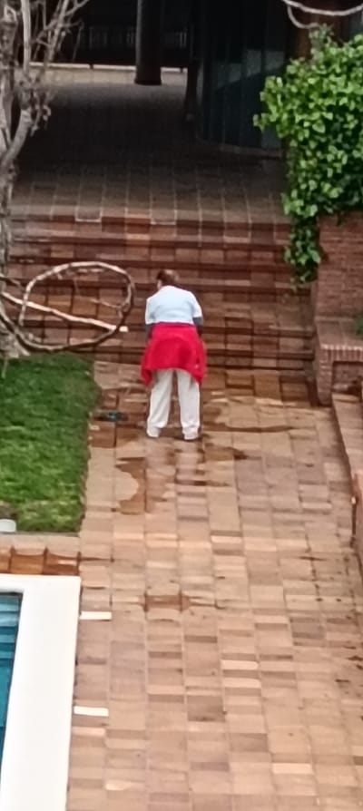 The image depicts an outdoor terraced area in Sant Cugat del Vallès, Spain. A single individual, seen from behind, is positioned centrally on a series of light brown/tan brick-paved steps. The person is wearing a light-colored, short-sleeved top and white pants, with a red garment tied around their waist. They are oriented away from the viewer, leaning slightly forward and downwards.

The environment consists of extensive light brown/tan rectangular brick paving, forming both the steps and flat ground surfaces. Numerous wet patches are visible on the lower paved areas, particularly on the flat surface in the mid-ground and foreground. To the immediate left of the steps is a strip of green grass, bordered by brick. Further left, a bare-branched tree or vine is present. To the right of the steps, a brick wall is topped with green foliage. In the lower left foreground, a section of a swimming pool with light blue water and white coping is visible. The background is darker, showing elevated paved areas and indistinct architectural elements, including what appear to be dark columns and a dark green wall section. No interactions are visible.