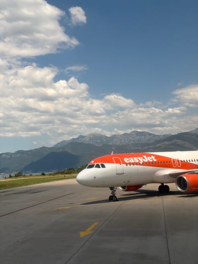 An EasyJet airplane is parked on a tarmac at an airport in Mrčevac, Montenegro. The plane is white with an orange stripe and the EasyJet logo on its side. The plane's engines are visible, as are its landing gear and wheels. In the background, there are mountains and a body of water. The sky is blue with white clouds. The tarmac is gray and cracked, with yellow markings and lines. The image is taken from a low angle, looking up at the plane.