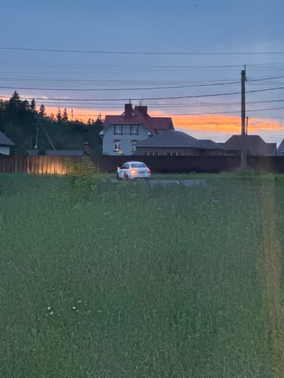 The image depicts a rural landscape at dusk in Chernushka, Russia. In the foreground, a field of tall green grass dominates the scene, with a few small white flowers scattered throughout. A wooden fence runs horizontally across the midground, separating the field from a residential area. Behind the fence, several houses with red tiled roofs and stone facades are visible. One house, with a prominent chimney, stands out. A white sedan with its headlights on is parked on a paved area near the houses. The sky above is a deep blue, transitioning to a vibrant orange and pink sunset. Power lines stretch across the sky, and a utility pole is visible on the right side of the frame. The overall impression is one of a quiet evening in a suburban or rural setting.
