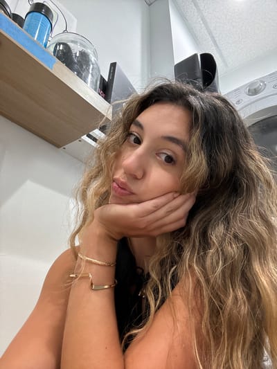 A young woman with long, wavy blonde hair rests her head on her hand, looking towards the camera. She is wearing a black top and a gold bracelet. In the background, a washing machine is visible, suggesting a laundry room or a home setting. To the left of the woman, a shelf holds several items, including a glass jar with hairbrushes inside, a blue hair roller, and a box labeled "KEUNE COLOR." The lighting in the image is bright and even, indicating it was likely taken indoors during the daytime. The woman's expression is soft and contemplative. The overall impression is that of a casual, personal moment captured in a domestic environment.
