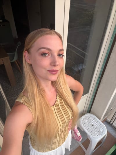 A young woman with blonde hair, light skin, and freckles poses for a selfie on a balcony. She is wearing a yellow, sleeveless, ruched top and a white skirt. Her right hand is on her hip, and her left arm is bent. A white plastic stool with a woven-pattern top is visible to her right. Behind her, a glass door shows a blurred view of an outdoor space. The scene appears to be in an urban setting, potentially in Catania, Italy, given the architectural style and outdoor elements.