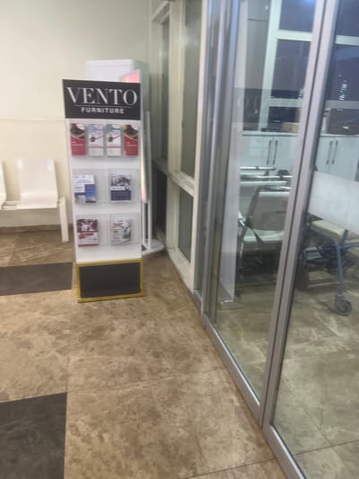 In the city of Karum, Nigeria, a retail display stand for "VENTO FURNITURE" stands prominently on a marble-tiled floor. The stand features multiple shelves showcasing various brochures and product images. Adjacent to the stand are two white chairs, suggesting a waiting area. To the right, a glass-paneled doorway offers a glimpse into another room, revealing several wheelchairs, some appearing to be in use. The lighting in the room is dim, with a bright light source visible in the background. The overall impression is one of a commercial space, possibly a furniture store or a showroom within a larger establishment.