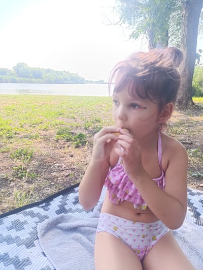 A young girl in a pink ruffled bikini with a yellow fruit pattern on her bottoms sits on a patterned blanket next to a grey towel. She is sitting outdoors on a grassy area with dirt and dry grass visible. The girl is eating something small held in her hands, and she is looking to her right with a thoughtful expression. Her brown hair is pulled up into a messy bun, and she has small pearl earrings. Behind her, a large tree stands to the right. In the background, a body of water, likely a river, is visible, with a tree-lined bank on the opposite side. The sun is bright, suggesting it is a warm, clear day, possibly midday or afternoon. The setting is a natural outdoor environment near a body of water, fitting for a recreational day in Zaliznyi, Russia. There is no visible text in the image.
