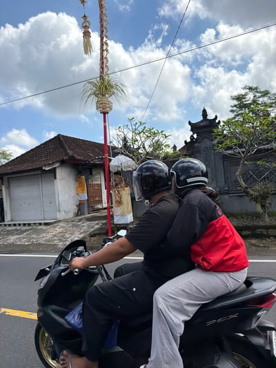 In Tampaksiring, Indonesia, two individuals are riding a black scooter on a paved road. The person in front is driving, wearing a black t-shirt and black pants, with a black helmet. The passenger behind them is wearing a red and black jacket and light grey pants, also with a black helmet, and is embracing the driver. The background shows a traditional Balinese building with a tiled roof and a decorative gate, as well as a tall red pole adorned with offerings and greenery. The sky is partly cloudy.