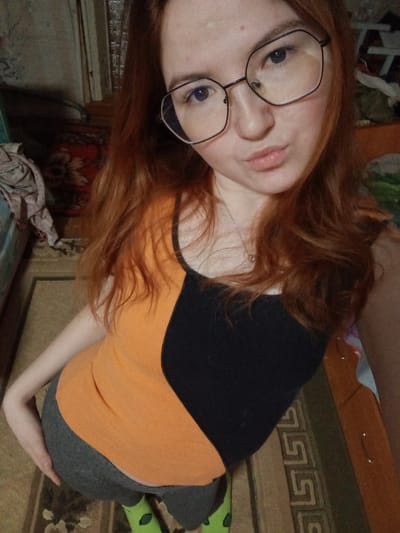 An indoor, high-angle portrait depicts a young female subject with long, reddish-brown hair, light-colored eyes, and facial blemishes, wearing dark-rimmed, hexagonal eyeglasses. She exhibits a neutral expression with slightly pouted lips and a visible silver chain with a small pendant around her neck. The subject is dressed in a sleeveless top, horizontally split into an orange upper section and a black lower section, grey form-fitting leggings, and bright green socks adorned with a repetitive pattern. Her left hand is positioned on her left hip or upper buttock. The background indicates a residential room in Старый Шагирт, Russia, featuring a patterned floor rug with geometric designs in beige, brown, and green tones. A dark red tapestry with a floral motif is mounted on a light-colored wall to the left, adjacent to a partially visible light blue furniture item draped with crumpled fabric. A wooden multi-drawer dresser is partially visible on the right side of the frame. The scene is illuminated by what appears to be ambient indoor lighting.