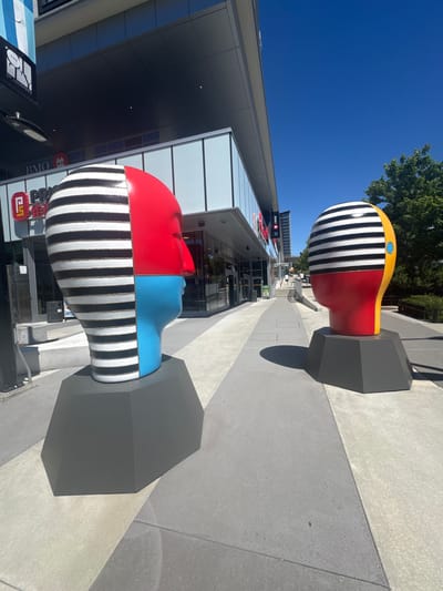Two large, stylized head sculptures are displayed on a paved outdoor plaza in front of a modern building in Burnaby, Canada. The sculpture on the left is closer to the camera and faces right, while the sculpture on the right is further away and faces left. Both sculptures depict the profile of a human head, divided into sections with different colors and patterns.

The left sculpture has its left side covered in black and white horizontal stripes. The right side of its face is divided into a red upper section and a blue lower section. It is mounted on a dark gray, faceted pedestal. Behind this sculpture, the building features large glass windows and signage that includes "BMO" and a red square logo with a stylized "P".

The sculpture on the right also features black and white horizontal stripes on its left side, and a yellow outer curve on its right. Its face is primarily red, with a small blue circular detail on the yellow section. It is also mounted on a dark gray, faceted pedestal. In the background, a tree with green foliage is visible against a clear blue sky. Several people are walking in the distance on the plaza. The ground is a light gray concrete with subtle tonal variations and intersecting lines.