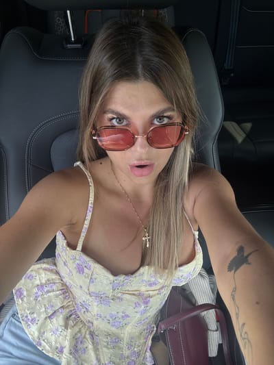 A young woman with blonde hair and sunglasses is taking a selfie in a car. She is wearing a yellow floral corset top and blue jeans. Her mouth is slightly open, and she is wearing a necklace with a cross pendant. There is a tattoo on her arm. The background shows the interior of a car, with black leather seats. The location is identified as a street in the city of Pastukhovo, Russia.