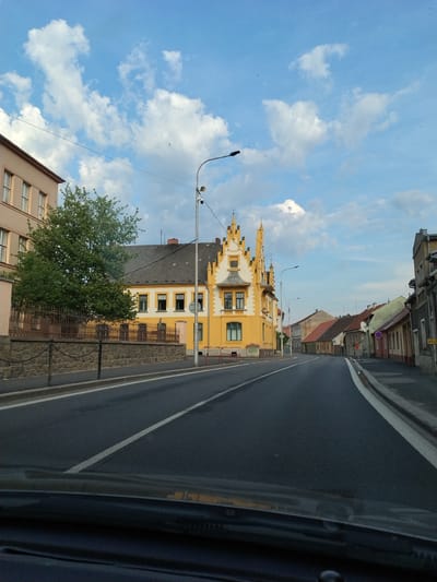The image shows a street view in Horažďovice, Czechia. The perspective is from a moving vehicle, indicated by the right side of the frame showing a car's interior. The scene is dominated by a bright yellow building with a distinctive ornate facade and a steep, pointed roofline. Adjacent to it is a darker building with a gray roof.
The street is paved and has white lane markings. A lamppost with lights and surveillance cameras stands near the yellow building. A metal fence runs along the side of the property, behind which is a stone wall. A large green tree with dense foliage is positioned in front of the fence and building. The sky is blue with scattered white, fluffy clouds, suggesting fair weather. Several small dark specks in the sky appear to be birds.
Other buildings line the street, some with red tile roofs, typical of European architecture. A red and white circular sign is visible on one of the buildings in the distance. The overall impression is a daytime scene in a European town or city. No visible text is legible in the image.