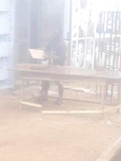 The image captures an outdoor scene in Enugu, Nigeria, characterized by a dusty, sandy ground and a general haze that softens details. In the mid-ground, a person, appearing to be male, stands behind a wooden table. The person is blurred but seems to be engaged in an activity involving the table. The table itself is a rustic wooden structure with a visible top and legs, set on the uneven ground. To the left of the table, a portion of a structure with lettering is visible, likely a building or a sign, with the partial word "NO" discernible. In the background, more structures and possibly some caged displays or shelves are present, also obscured by the atmospheric haze and low image quality. The overall impression is of a marketplace or a street-side stall.