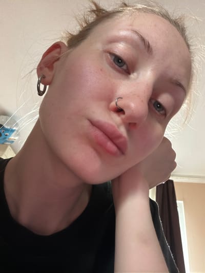 The image is a close-up, tilted selfie of a young woman with blonde hair. She is wearing a black top and has a silver hoop earring in her left ear and a silver septum piercing. Her right arm is bent and resting near her face, with her hand touching her cheek. Her eyes are a light blue, and her lips are slightly parted. She has a natural makeup look, with a few freckles visible on her nose and cheeks. The background appears to be indoors, with a wall and a curtain visible. The lighting suggests it could be daytime. There is no visible text in the image. The context provided suggests the location is Ladozhskoe Ozero, Russia.