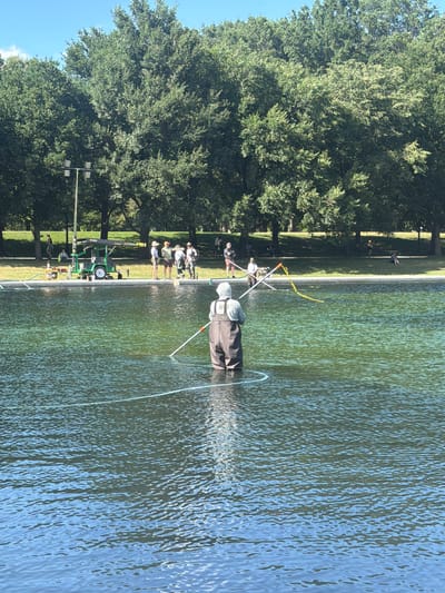 The image shows a sunny day at the Tidal Basin in Washington, D.C. A person wearing waders and a hooded jacket is standing in the water, holding a long pole with a yellow streamer attached to it. The person appears to be working in the water, possibly cleaning or inspecting it. In the background, several other people are gathered on the grassy bank, some of whom appear to be observing the person in the water. There is a small green utility vehicle with a crane on the side, and some equipment is visible near the edge of the basin. Tall trees line the background, and the sky is clear and blue. The water in the Tidal Basin is a greenish-blue color.