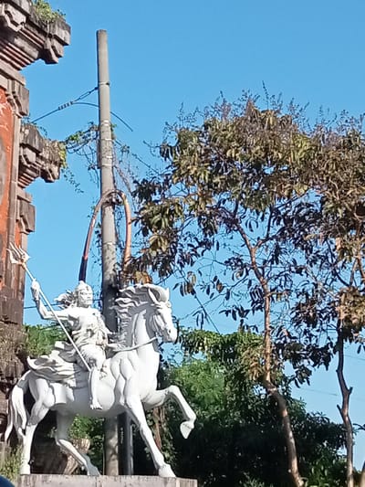 This image depicts a white sculpture of a person riding a horse, positioned in front of a rustic brick structure with decorative stonework. The sculpture is in a dynamic pose, with the horse rearing and the rider holding a trident aloft. The figure on the horse appears to be a male, with flowing hair and a determined expression. The overall style of the sculpture suggests it might be of religious or historical significance.
The background is dominated by a clear blue sky and lush green trees, some of which have visible fruit or seed pods. A utility pole with wires runs vertically behind the sculpture, adding a modern element to the scene. The lighting suggests it is daytime, with the sun casting shadows and highlighting the details of the sculpture and the surrounding foliage. The setting appears to be outdoors, possibly in a temple complex or a public space in Blahbatuh, Indonesia, given the architectural style of the brick structure and the tropical vegetation. There are no visible signs of any specific activity or event, other than the static presentation of the sculpture itself. The atmosphere is serene and bright, with the blue sky and green trees contributing to a sense of natural beauty and tranquility.