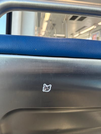 The image displays the interior of a public transit vehicle, identified as a Metrorail train car, situated within Hialeah, United States of America. In the immediate foreground, a segment of a royal blue, textured vinyl cushion, likely part of a seat or barrier, is visible. Below this cushion is a brushed metallic panel. Centered on this metallic surface, slightly to the left, is a small, die-cut sticker, approximately 3 centimeters in width. The sticker depicts the head of a gray cat wearing black, pixelated sunglasses and holding a small, white, cylindrical object in its mouth. The metallic panel exhibits minor scuff marks. The background, rendered out of focus, reveals the train car's ceiling, overhead fluorescent strip lighting, and multiple metallic grab rails and straps, consistent with public transportation infrastructure. No individuals are present or discernable.