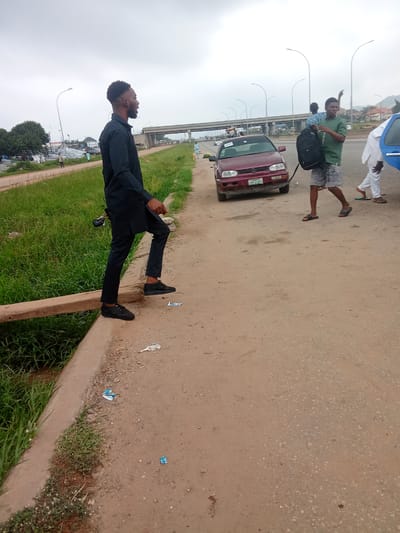 In Kagini, Nigeria, on a cloudy day, a young man in a black outfit stands on the edge of a dirt road, his right foot on a wooden plank that extends over a grassy area. He is facing left, with his mouth slightly open, as if speaking. Behind him, a red Volkswagen Golf car is parked on the road. To the right of the car, another young man in a green t-shirt and shorts, carrying a black backpack, walks away from the camera. Further to the right, a person in white clothing is partially visible, seemingly bending down near the open door of a blue car. In the background, an overpass looms against the overcast sky, with more cars and distant buildings visible. The scene depicts a roadside environment with evidence of human activity and vehicles.