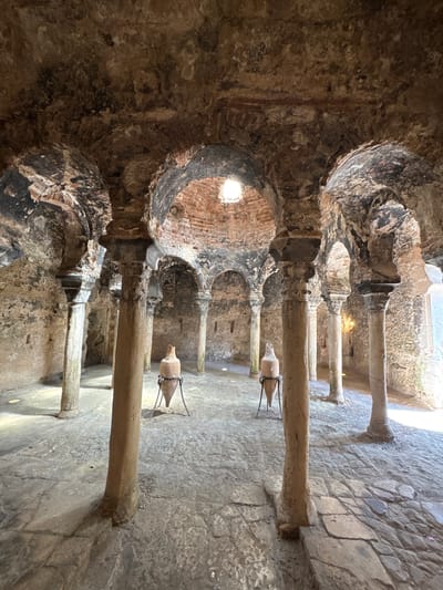 This is a wide-angle, eye-level photograph of the interior of the historic Arab Baths (Baños Árabes) located in Palma de Mallorca, Spain. The scene depicts a vaulted chamber with rough, textured stone walls and ceiling, exhibiting signs of age and wear. Several stone columns, some with intricately carved capitals, support the arched ceiling. Light filters in from a circular opening in the ceiling and from an opening on the right side of the frame. Two large, antique clay amphorae, each supported by a metal stand, are placed on the stone-paved floor. The floor itself is uneven and composed of various sizes of stone slabs. The overall atmosphere is one of historical preservation and antiquity.