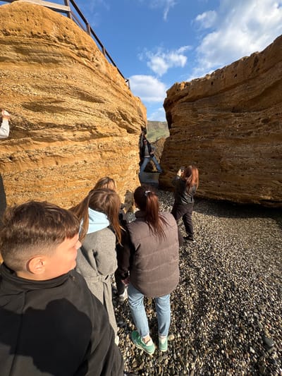 The image captures a group of people lying on a pebble beach beneath a large rock formation in Novyi Svet, Ukraine. The sky is visible with scattered clouds. A wooden and metal railing can be seen along the upper edge of the lower rock formation. One person is taking a photograph of the upper rock formation with a mobile phone.