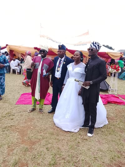 In an outdoor setting in Kuru, Nigeria, a wedding party is in progress. The bride, in a long white gown, stands next to a man in a black outfit, holding a microphone and a piece of paper. To the left of the bride stands a man in a military-style uniform and hat adorned with medals. To the far left is a man dressed in a maroon robe and green patterned pants, holding a bucket. A crowd of people sits under a tent adorned with red and gold fabric in the background. A small table holding flowers sits to the right. Two pink carpets are on the ground.