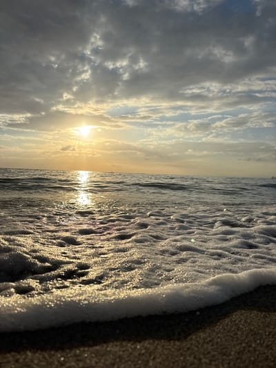 The image depicts a low-angle view of a beach at sunrise. The sun is low on the horizon, casting a warm, golden light across the scene. The sky is partly cloudy, with dramatic, textured clouds illuminated by the sun. The ocean is calm, with gentle waves lapping at the shore. The foreground is dominated by foamy white water and wet sand, reflecting the light from the sun and the sky. The image evokes a sense of tranquility and natural beauty. The location is the beach in Abang, Bali, Indonesia.