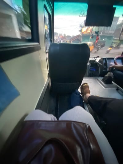 The image depicts the interior front of a bus, looking out onto a street in Jatisari, Indonesia. In the foreground, the lower body of an individual, wearing light-colored, possibly ribbed pants, is visible with a dark brown bag resting on their lap. Directly ahead is an unoccupied dark-colored passenger seat. To the right, the lower legs and right foot of another person, shod in a brown sandal, are visible near the vehicle's dashboard and controls, indicating a driver or front passenger. A blue bag is on the floor beside the unoccupied seat. Outside, a street scene reveals multiple motorcycles, a yellow three-wheeled vehicle resembling an auto-rickshaw or becak, and various single to two-story buildings, some with visible signage. Overhead utility lines crisscross the sky. The ambient light suggests daytime conditions.