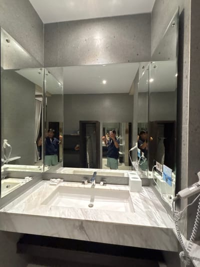 A person is seen taking a selfie in a modern bathroom located in Batuan, Indonesia. The image shows a double vanity with two rectangular white sinks integrated into a light-colored, veined marble countertop. Above the vanity is a large, segmented mirror consisting of multiple rectangular panels, creating a multi-faceted reflection. The walls surrounding the mirror are a textured grey.

The individual, wearing a dark blue polo shirt and light-colored shorts, is reflected prominently in the main mirror, holding a mobile phone to capture the image. Their reflection is repeated and slightly distorted across the angled mirror panels.

On the left side of the vanity, a small rectangular tray holds two round white objects. In the center, a white rectangular holder with three compartments is visible. On the right side, a white hairdryer with a coiled cord is mounted on the wall or resting on the counter, next to a holder displaying small blue cards. Through the mirror's reflection, a white curtain is visible to the left, and an open dark wood wardrobe or shelving unit, along with an ironing board and iron, can be seen in the background to the right. The ceiling is white with visible recessed lighting fixtures.