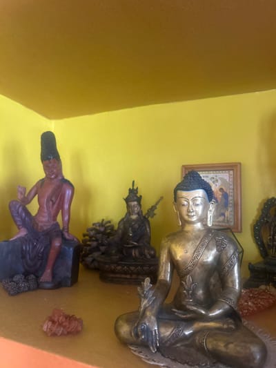 The image shows a shelf in a room, likely in a house, with a yellow wall. On the shelf are several statues. A brass-colored statue of a seated Buddha is prominent. Below the Buddha are other statues, including one of a dark-skinned male figure with a distinctive hairstyle. A framed picture hangs on the wall behind the Buddha statue. Additionally, there is a small cluster of reddish-brown mineral formations. The specific location in Puttaparthi, India is not discernible from the image.