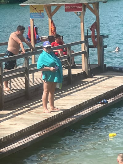 This image depicts a sunny day at a lake or waterfront in Vilnius, Lithuania. The scene is centered around a wooden pier extending into clear blue water. Several people are present on and around the pier. In the background, a person is swimming in the water. On the pier, a person wearing a blue hat and a teal cover-up is holding a phone. Another person is climbing a ladder on the pier, and a third person is shirtless, wearing patterned shorts, and appears to be resting or preparing to enter the water.
Objects visible include a life preserver hanging on the pier structure, a paddleboard near the edge of the pier, and two yellow buoys floating in the water. The pier itself is constructed of light-colored wooden planks, casting long shadows that indicate it is likely midday or early afternoon. The water is calm with gentle ripples.
There is visible text on a red sign affixed to the pier structure that reads "GELBĖTOJAI", which translates to "Lifeguards" in Lithuanian. Below this, another sign with yellow and blue elements displays a pictogram of a person diving and the text "DRAUDŽIAMA NARDYTI", meaning "Diving prohibited". The overall atmosphere seems relaxed and recreational, with people enjoying a day by the water.