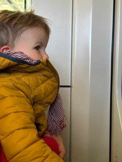 A close-up, horizontal shot features a young child, likely a toddler or preschooler, primarily from the chest up. The child has light brown hair and is wearing a mustard yellow quilted puffer jacket and a red and white striped scarf or blanket around their neck. Their eyes are cast upwards and slightly to the left, indicating they are looking at something out of frame.

The background consists of light grey or off-white paneled surfaces, suggesting the interior of a vehicle, possibly a train or bus, which aligns with the location context of Ostend, Belgium, where public transport is common. A glimpse of a window with soft, possibly green or yellow, light reflection is visible behind the child's head. Part of an adult's hand, wearing red nail polish and resting on a red surface (possibly another piece of clothing or a cushion), is visible to the right of the child, implying they are being held or accompanied. The overall impression is one of quiet observation or travel. There is no visible text in the image.