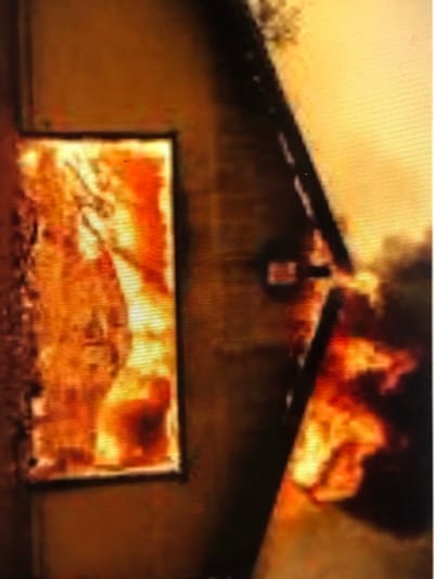 This image depicts a structure in Kafr Sousa Municipality, Syria, actively engulfed in fire. On the left side, a rectangular opening, consistent with a window or framed aperture, is filled entirely with intense orange and yellow flames, indicating an internal blaze. To the right, a darker structural element, possibly a wall or beam, is visible, with more flames and dark smoke billowing from beneath and around it. A diagonal dark line, likely representing a roofline or architectural feature, separates areas of fire and smoke. The scene is dominated by the bright, luminous glow of the fire, casting warm tones across the visible surfaces, with no discernible individuals or explicit actions beyond the ongoing combustion.