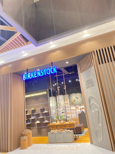 The image shows an indoor retail environment, rotated 90 degrees clockwise, displaying the entrance and interior of a Birkenstock store. The perspective is looking down into the store from an upper level or a high vantage point.

A bright blue neon sign prominently displays the brand name "BIRKENSTOCK" horizontally, marking the entrance. The store interior features modern design elements, including light wood or wood-paneled walls and ceiling accents. One wall inside the store is adorned with a large, embossed white shape resembling a footbed, flanked by the text "GERMAN TRADITION SINCE 1774". The same text appears vertically on another darker wall.

The store is well-lit and appears organized. Various Birkenstock sandals and shoes are displayed on shelves and wooden stands along the walls. Several display units, some with a concrete-like finish and wooden drawers, showcase merchandise. One display unit also bears the "BIRKENSTOCK" text. Decorative ropes with knots are strung across the store, adding to the visual appeal. A small green plant sits on a display table. The flooring inside the store seems to be a combination of light wood and darker sections. No people are visible, and no specific activity is taking place, suggesting the store is open for business. The setting is likely a shopping mall or a commercial building in Bekasi Regency, Indonesia.