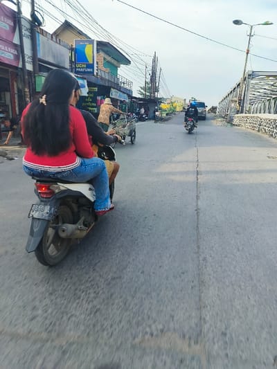 The image shows a scene from Baleendah, Indonesia (latitude: -6.992, longitude: 107.626), a woman wearing a red shirt and blue jeans is riding a motorcycle with a male passenger in the rear. The motorcycle is moving towards the camera. There is a single lane of traffic, and the woman is looking straight ahead. The motorcycle is passing a small truck, which is parked on the right-hand side of the road. There is another motorcycle in the distance going in the opposite direction. There are several small shops and buildings in the background on the left side of the road, a metal bridge spanning a river in the background, and a gray, paved road in the foreground. The sky is mostly cloudy and gray.  The atmosphere is quiet and uneventful.