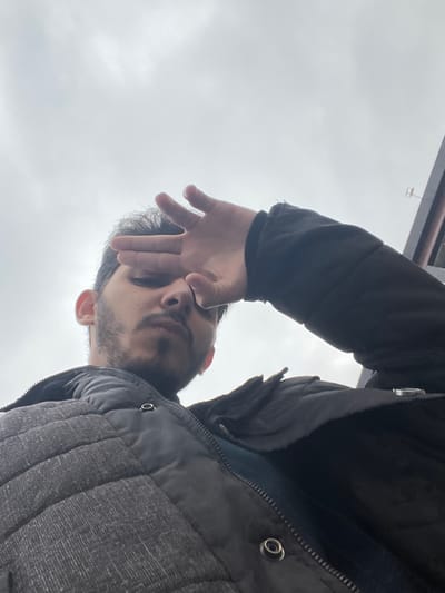 A low-angle outdoor photograph depicts a male individual, appearing to be a young adult, with short dark hair, a beard, and a mustache. He is wearing a dark grey quilted puffer vest over a dark long-sleeved garment. His right hand is raised with fingers slightly splayed, palm covering his forehead and eyes, shielding them. The background is dominated by a bright, overcast light grey sky. In the upper right portion of the image, a dark, angled structural element, consistent with a building facade or roofline, is partially visible, featuring a faint, light-colored 'H' shaped fixture. The scene is located in Al-Mazza Municipality, Syria.