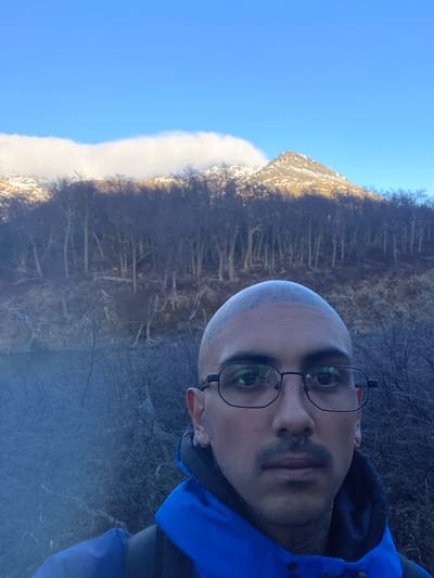 A selfie taken by a person with a shaved head and glasses wearing a blue jacket. Behind the person is a view of Iguazu Falls, located in Unknown, Argentina. The sky is blue.