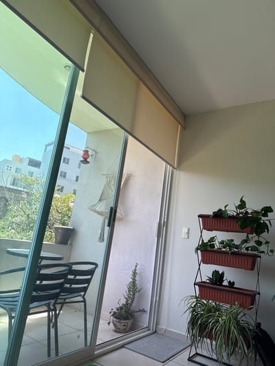 The image, oriented vertically, captures a view from an indoor space looking out onto an outdoor balcony. The scene is brightly lit by natural daylight, suggesting it is daytime with clear weather.

Indoors, the walls are painted a light, neutral color. On the right side, a wall-mounted metal plant stand holds three rectangular brown planters. These planters house various green plants, including what appears to be a pothos vine, a snake plant, and a lush spider plant with striped leaves. Below the plant stand, a white light switch is visible on the wall. Over the large sliding glass door or window that leads to the balcony, several light-colored (beige/tan) roller blinds are partially or fully rolled up. A thin gray mat or rug is visible on the floor near the doorway on the right.

Through the clear glass of the sliding door, the outdoor balcony is visible. The balcony features light-colored tiled flooring. A white macrame hammock is artfully hung on the balcony's wall. A red and blue bird feeder or decorative item is also attached to the wall. Further out on the balcony, a small black metal outdoor table is visible, along with two matching black metal chairs, which are partially stacked.

Beyond the balcony, an urban or suburban landscape unfolds under a bright blue sky. Several light-colored buildings with multiple rectangular windows are visible, along with abundant green trees. The overall impression is a cozy living space connected to a balcony, offering a view of a residential area, likely in Cuernavaca, Mexico. There are no people visible, and no discernible text.