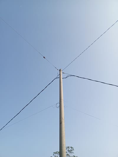 A low-angle shot focuses on a concrete utility pole in Thalang, Laos, against a pale blue sky. The pole is centrally positioned and extends vertically, intersected by several power lines that radiate outwards, forming a crisscross pattern. One line has a small, dark object attached. A crescent moon is visible in the upper left of the frame. At the base of the pole, a glimpse of trees or foliage is visible.